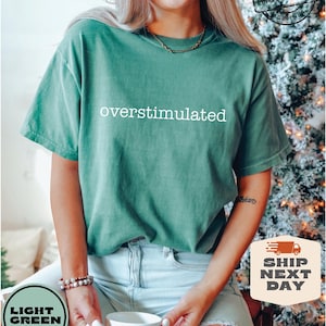 May include: Light green t-shirt with the word "overstimulated" in white. The shirt is made of Comfort Colors 100% cotton. The model is wearing light blue jeans and holding a white mug. The shirt is labeled "Light Green" and "Ship Next Day".