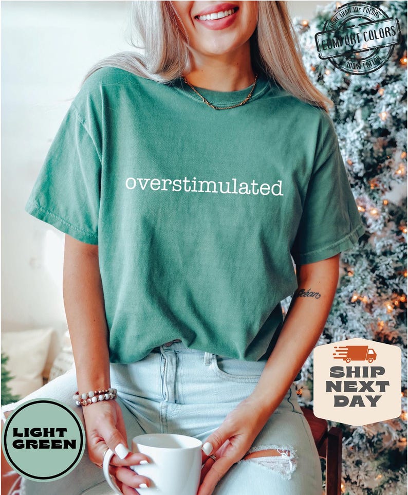 May include: Light green t-shirt with the word "overstimulated" in white. The shirt is made of Comfort Colors 100% cotton. The model is wearing light blue jeans and holding a white mug. The shirt is labeled "Light Green" and "Ship Next Day".