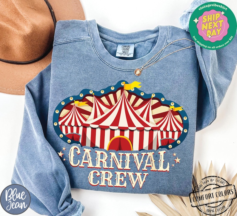 May include: A blue crew neck sweatshirt with a carnival tent graphic in red and white stripes. The text "CARNIVAL CREW" is printed below the graphic in a vintage style font. The sweatshirt has a relaxed fit.