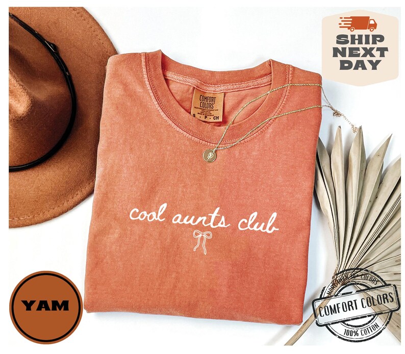 May include: A peach-colored t-shirt with the text "cool aunts club" in white lettering. The shirt has a distressed look and is made of 100% cotton.