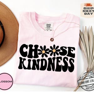 May include: Pink t-shirt with the text "Choose Kindness" in a retro font with three yellow flowers. The shirt is made of 100% cotton.