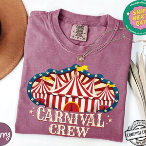 May include: Berry-colored t-shirt with a circus tent graphic and the words "CARNIVAL CREW". The graphic features red and white striped tents with blue and yellow accents. The shirt has a crew neck and short sleeves.