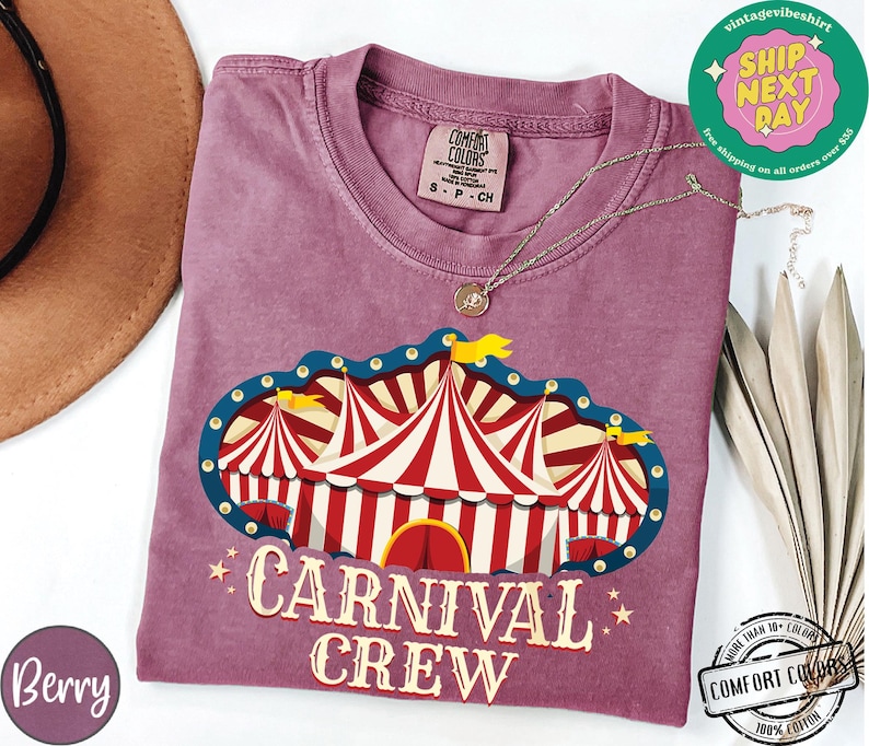 May include: Berry-colored t-shirt with a circus tent graphic and the words "CARNIVAL CREW". The graphic features red and white striped tents with blue and yellow accents. The shirt has a crew neck and short sleeves.