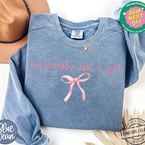 Puede incluir: Una sudadera de mezclilla azul con el texto "I'm literally just a girl" en letras rosas. Un lazo rosa est&aacute; centrado debajo del texto.