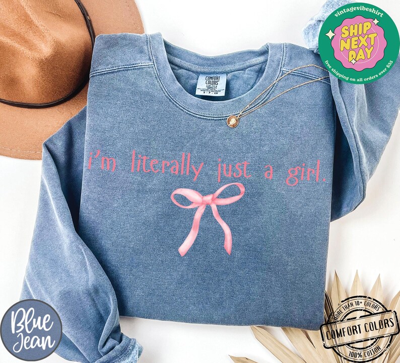 Puede incluir: Una sudadera de mezclilla azul con el texto "I'm literally just a girl" en letras rosas. Un lazo rosa est&aacute; centrado debajo del texto.