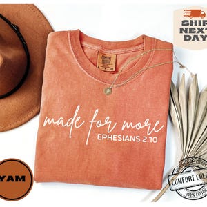 Puede incluir: Una camiseta de color coral con el texto "made for more" y "EPHESIANS 2:10" en blanco. La camiseta est&aacute; hecha de 100% algod&oacute;n.