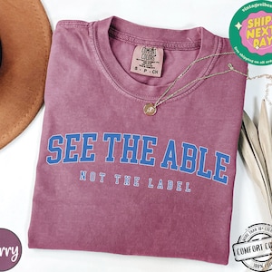 May include: A maroon t-shirt with the text "See the Able, Not the Label" in blue lettering. The shirt is made of a soft, comfortable fabric.