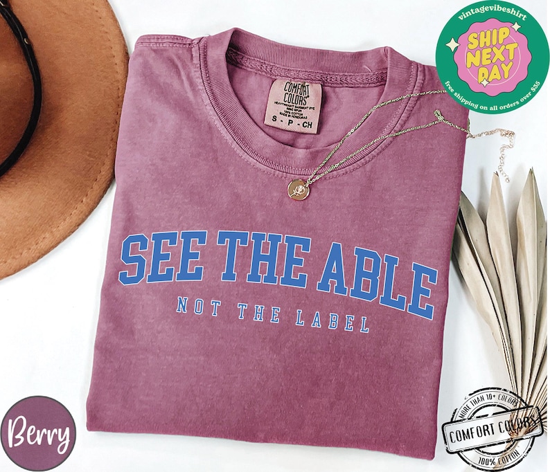 May include: A maroon t-shirt with the text "See the Able, Not the Label" in blue lettering. The shirt is made of a soft, comfortable fabric.