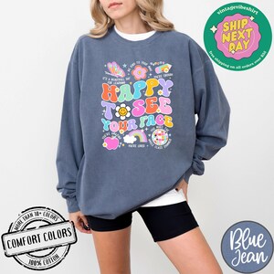 May include: A dark blue sweatshirt with a colorful graphic design that says "Happy to see your face". The graphic includes a rainbow, a butterfly, a flower, a smiley face, and a disco ball. The sweatshirt is made of 100% cotton.