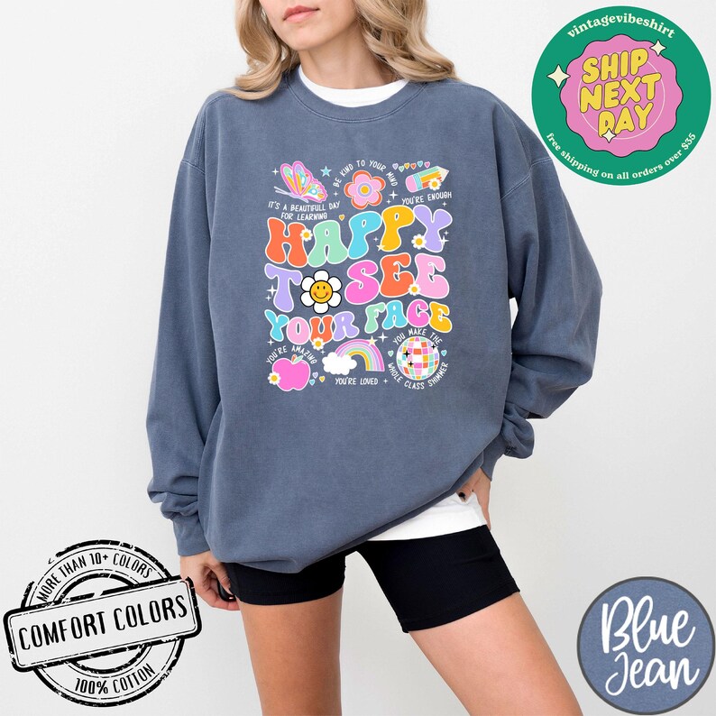 May include: A dark blue sweatshirt with a colorful graphic design that says "Happy to see your face". The graphic includes a rainbow, a butterfly, a flower, a smiley face, and a disco ball. The sweatshirt is made of 100% cotton.