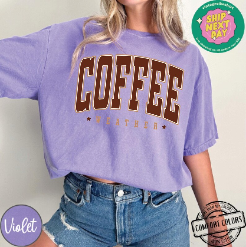 May include: A purple t-shirt with the text "COFFEE WEATHER" in brown letters with a white outline. The shirt is made of a soft, comfortable material.