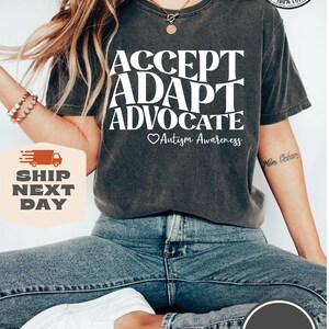 May include: A dark gray t-shirt with the text "ACCEPT ADAPT ADVOCATE" in white, with a heart and the words "Autism Awareness" below. The model is wearing blue jeans and white sneakers.