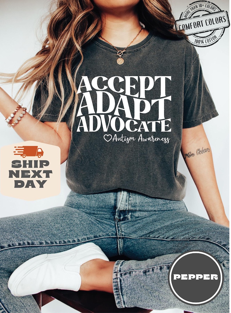 May include: A dark gray t-shirt with the text "ACCEPT ADAPT ADVOCATE" in white, with a heart and the words "Autism Awareness" below. The model is wearing blue jeans and white sneakers.