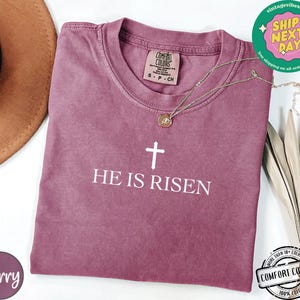 May include: A mauve colored t-shirt with a white cross and the text "HE IS RISEN".