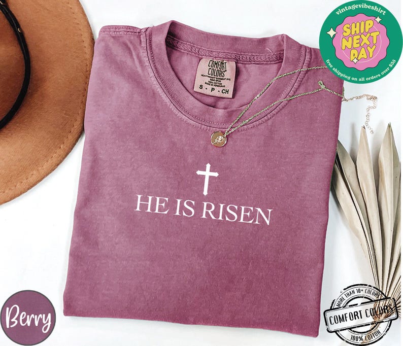 May include: A mauve colored t-shirt with a white cross and the text "HE IS RISEN".