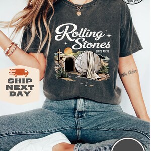 May include: A gray t-shirt with a graphic of a stone rolling away from a tomb. The graphic includes a cross, a sunrise, and the text "Rolling Stones Since AD 33".
