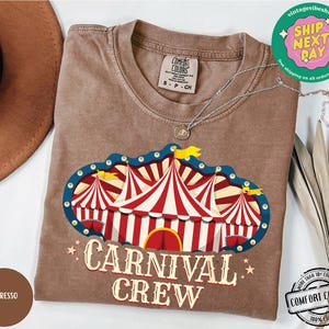 May include: A brown t-shirt with a circus tent graphic in red, white, and blue, with the words "CARNIVAL CREW" in a vintage font. The shirt is made of Comfort Colors 100% cotton.