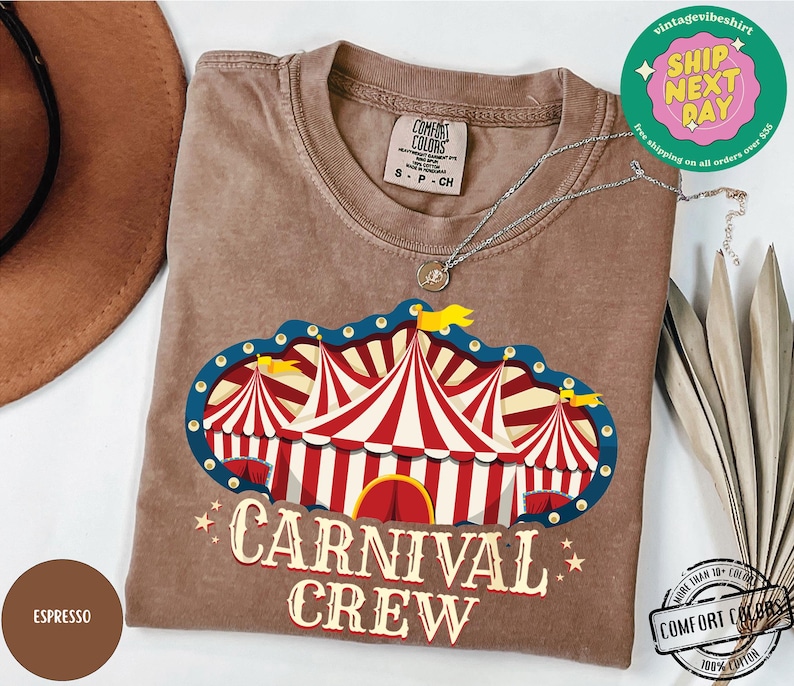 May include: A brown t-shirt with a circus tent graphic in red, white, and blue, with the words "CARNIVAL CREW" in a vintage font. The shirt is made of Comfort Colors 100% cotton.