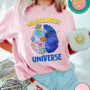 Op de afbeelding: Roze T-shirt met een afbeelding van een brein met planeten en sterren erin. De tekst "Neurodiverse Universe" staat op het shirt gedrukt.
