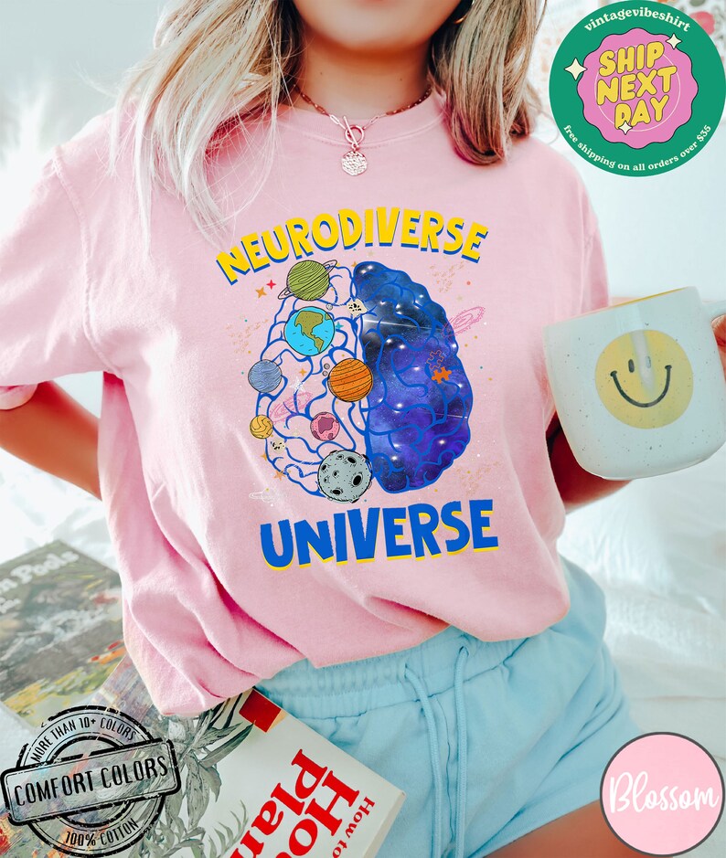 Op de afbeelding: Roze T-shirt met een afbeelding van een brein met planeten en sterren erin. De tekst "Neurodiverse Universe" staat op het shirt gedrukt.