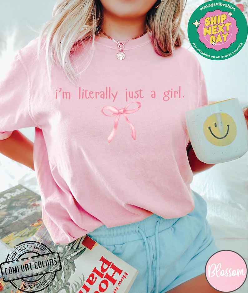 Puede incluir: Una camiseta rosa con el texto "i'm literally just a girl." y un lazo rosa. La camiseta la lleva puesta una persona con el pelo rubio y pantalones cortos azules.