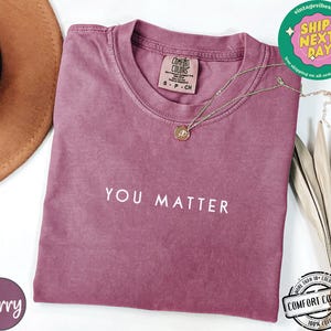 May include: A mauve colored t-shirt with the text "YOU MATTER" printed in white on the front. The shirt is made of a soft, comfortable fabric and has a vintage look.