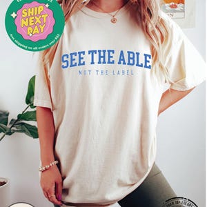 May include: A white t-shirt with the text "See the Able, Not the Label" in blue lettering. The shirt is being modelled by a person wearing black biker shorts.