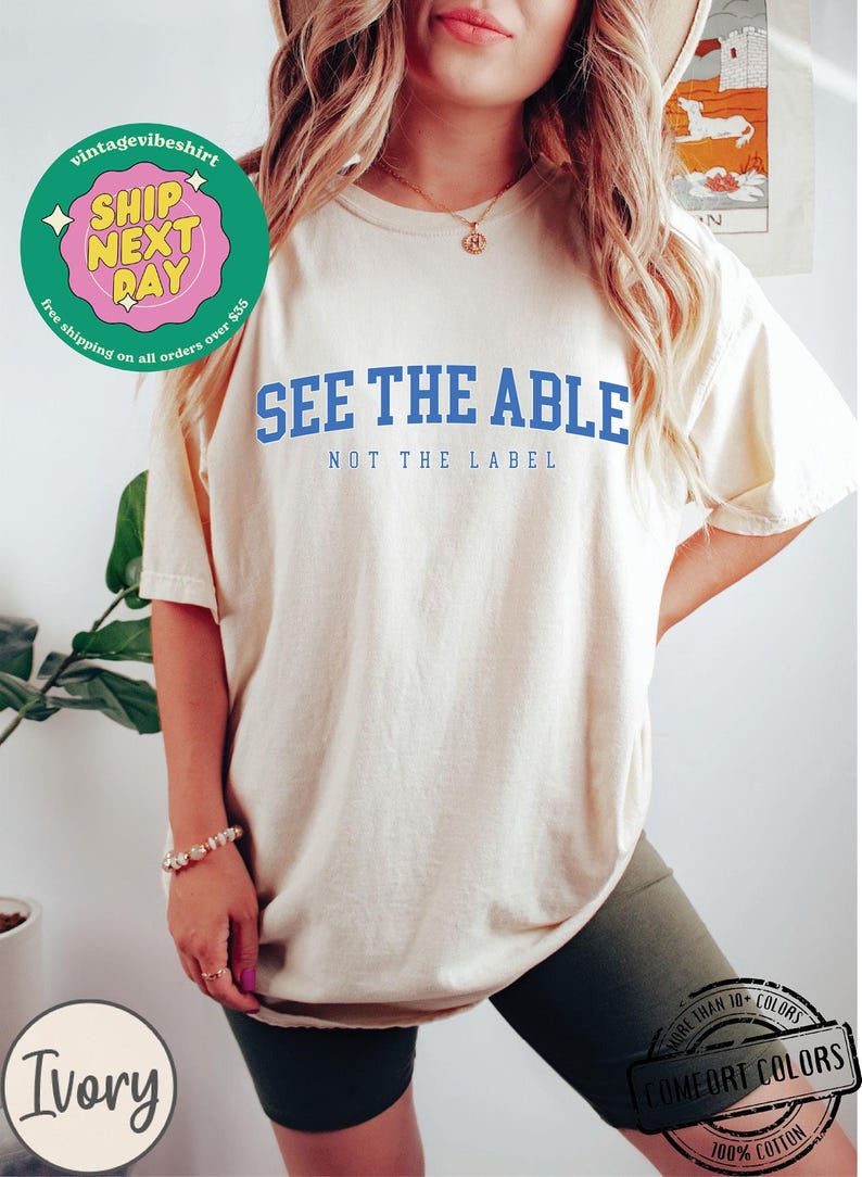 May include: A white t-shirt with the text "See the Able, Not the Label" in blue lettering. The shirt is being modelled by a person wearing black biker shorts.