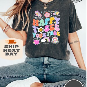 May include: A dark grey t-shirt with a colorful graphic that says "Happy to see your face". The graphic includes a butterfly, a flower, a rainbow, a disco ball, and a heart. The shirt is made of 100% cotton and is available in multiple colors.