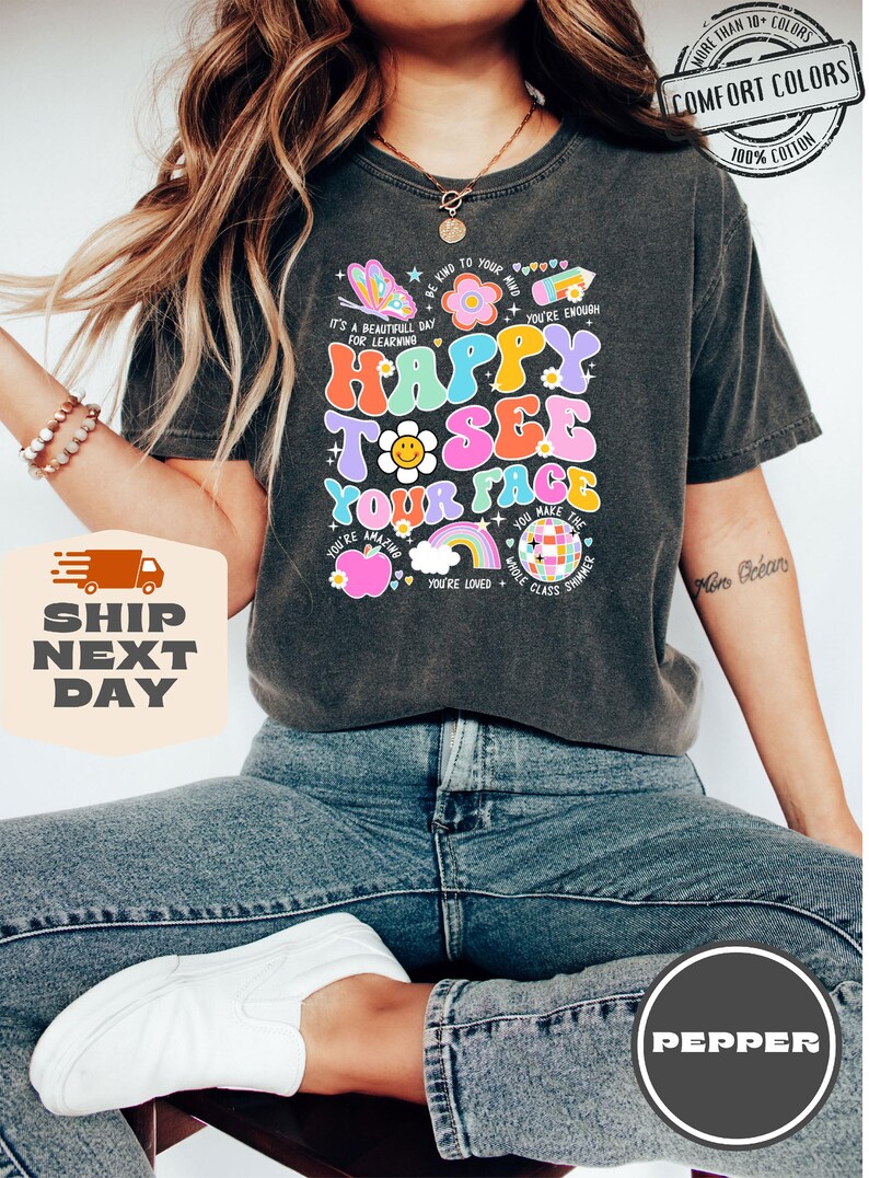 May include: A dark grey t-shirt with a colorful graphic that says "Happy to see your face". The graphic includes a butterfly, a flower, a rainbow, a disco ball, and a heart. The shirt is made of 100% cotton and is available in multiple colors.