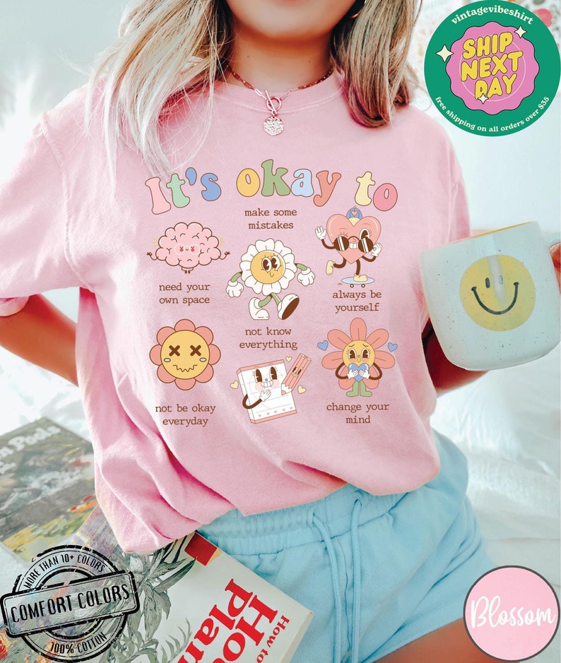 May include: Pink t-shirt with the text "It's okay to" and illustrations of various cartoon characters. The text below each character includes phrases like "make some mistakes", "need your own space", "not know everything", "always be yourself", "not be okay everyday", and "change your mind".