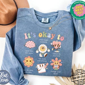 May include: A light blue sweatshirt with the text "It's okay to" and a series of colorful illustrations and text. The illustrations include a brain, a flower, a heart with sunglasses, a flower with a face, a notebook, and a flower with a face. The text includes "make some mistakes", "need your own space", "not know everything", "always be yourself", "not be okay everyday", and "change your mind".