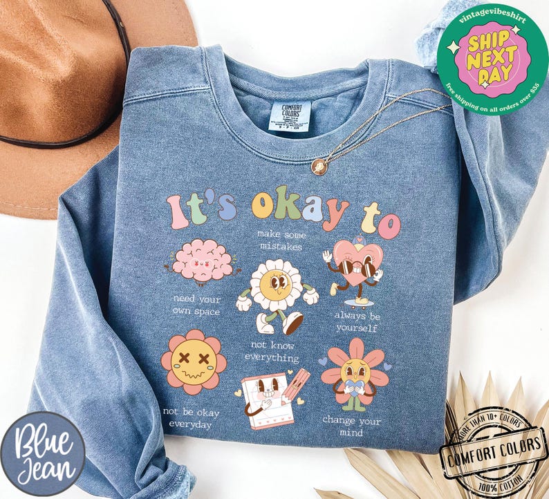 May include: A light blue sweatshirt with the text "It's okay to" and a series of colorful illustrations and text. The illustrations include a brain, a flower, a heart with sunglasses, a flower with a face, a notebook, and a flower with a face. The text includes "make some mistakes", "need your own space", "not know everything", "always be yourself", "not be okay everyday", and "change your mind".