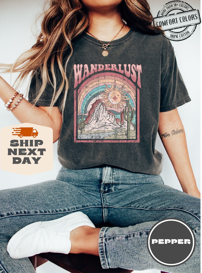 May include: Dark gray Comfort Colors t-shirt with a desert-themed graphic. The design features a rainbow, sun, mountains, and cacti, with the word "WANDERLUST" above. The shirt is 100% cotton and available in more than 10 colors.
