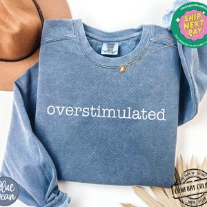May include: A blue crew neck sweatshirt with the word "overstimulated" embroidered in white. The sweatshirt has a vintage look and feel. A gold necklace is draped across the front. The sweatshirt is made of 100% cotton.