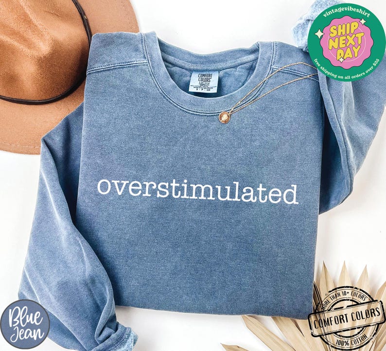 May include: A blue crew neck sweatshirt with the word "overstimulated" embroidered in white. The sweatshirt has a vintage look and feel. A gold necklace is draped across the front. The sweatshirt is made of 100% cotton.