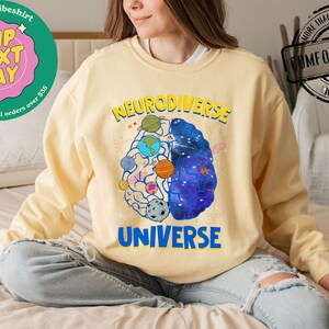 Op de afbeelding: Een geel sweatshirt met een afbeelding van een brein met planeten en sterren erin. De tekst "Neurodiverse Universe" staat op het sweatshirt gedrukt.