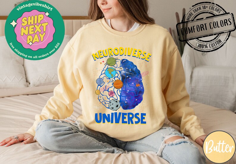 Op de afbeelding: Een geel sweatshirt met een afbeelding van een brein met planeten en sterren erin. De tekst "Neurodiverse Universe" staat op het sweatshirt gedrukt.