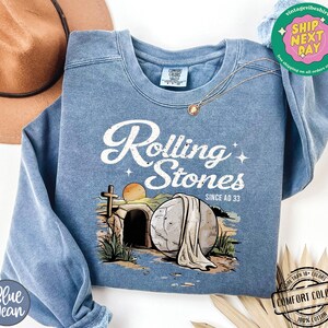 May include: A blue sweatshirt with a graphic of a stone tomb with a white cloth draped over it. The text "Rolling Stones Since AD 33" is printed above the image.