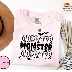 May include: Pink t-shirt with the text "MOMSTER" repeated three times in black dripping letters. The shirt has a spider and bats graphic. The text "COMFORT COLORS" is visible on the tag.