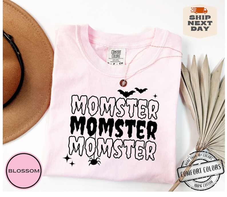 May include: Pink t-shirt with the text "MOMSTER" repeated three times in black dripping letters. The shirt has a spider and bats graphic. The text "COMFORT COLORS" is visible on the tag.