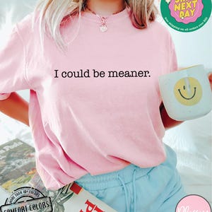 May include: A light pink Comfort Colors t-shirt with the text "I could be meaner." A white mug with a yellow smiley face is held in the hand. The image also shows a book and blue shorts.