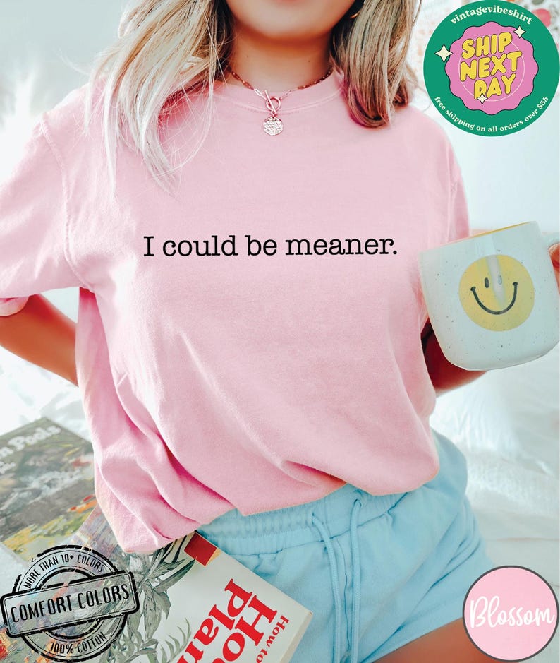 May include: A light pink Comfort Colors t-shirt with the text "I could be meaner." A white mug with a yellow smiley face is held in the hand. The image also shows a book and blue shorts.