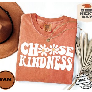 May include: A peach-colored t-shirt with the words "Choose Kindness" in white with a daisy graphic. The shirt is made of 100% cotton.