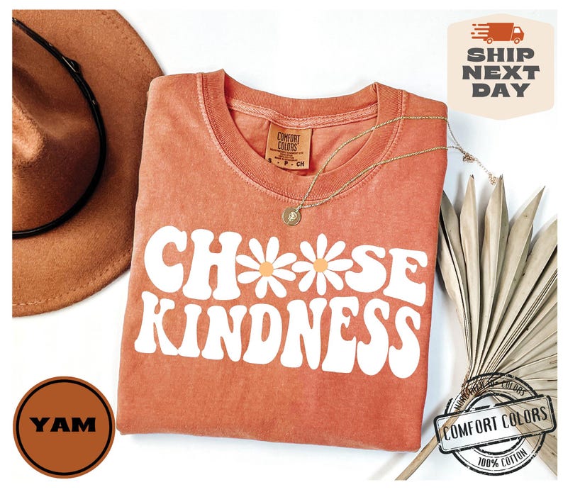 May include: A peach-colored t-shirt with the words "Choose Kindness" in white with a daisy graphic. The shirt is made of 100% cotton.