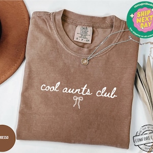 May include: A brown t-shirt with the text "cool aunts club" in white lettering with a white bow underneath. The shirt is made of a soft, comfortable fabric.