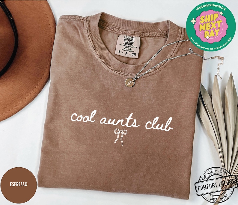 May include: A brown t-shirt with the text "cool aunts club" in white lettering with a white bow underneath. The shirt is made of a soft, comfortable fabric.