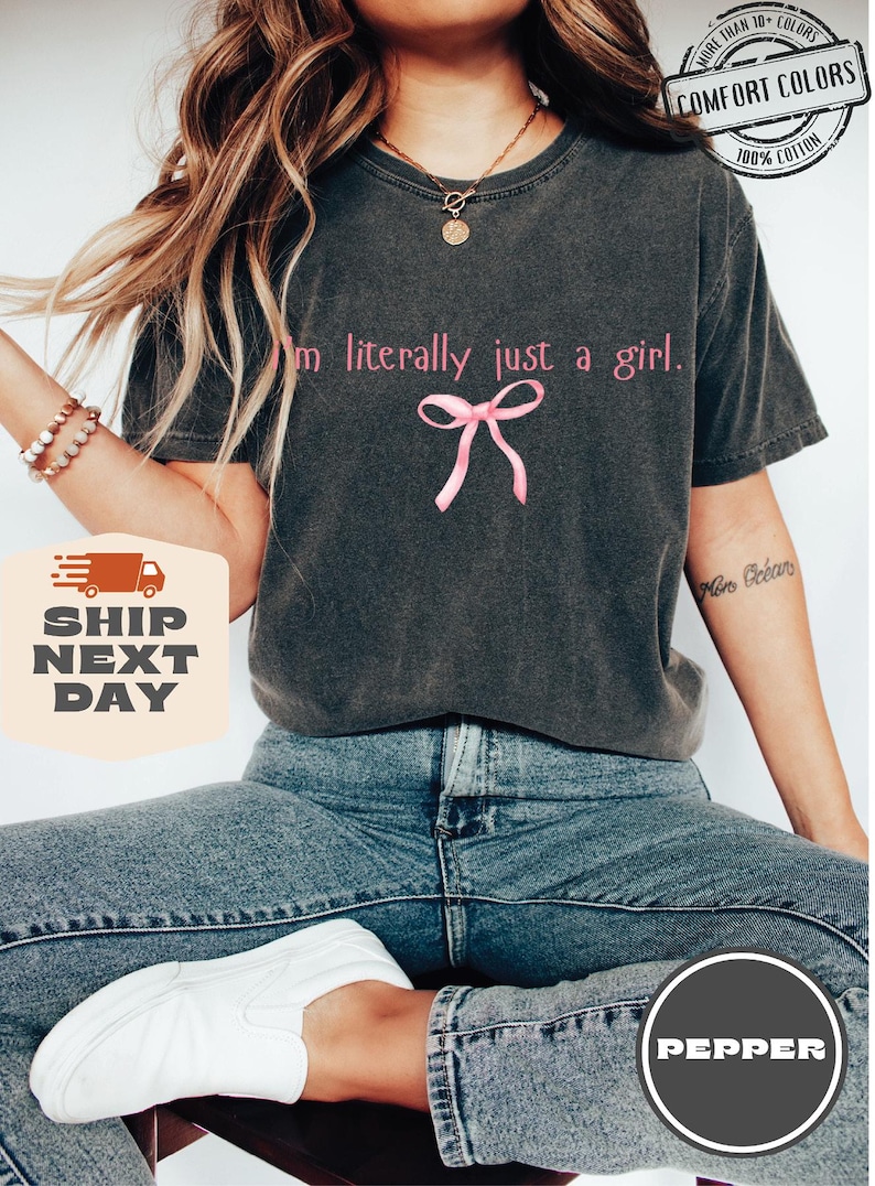 Puede incluir: Una camiseta gris oscuro con el texto "Soy literalmente solo una chica." y un lazo rosa. La camiseta est&aacute; hecha de 100% algod&oacute;n y est&aacute; disponible en varios colores.
