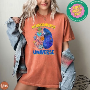 Op de afbeelding: Een oranje T-shirt met een afbeelding van een brein met planeten en sterren erin. De tekst "Neurodiverse Universe" is op het shirt gedrukt.