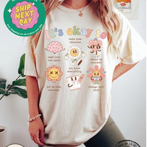 May include: A cream-colored t-shirt with the text "It's okay to" and a series of colorful illustrations of cartoon characters with positive messages. The illustrations include a brain, a flower, a heart, a flower with a sad face, a calendar, and a flower with a heart. The text below each illustration includes phrases like "make some mistakes", "need your own space", "not know everything", "always be yourself", "not be okay everyday", and "change your mind".
